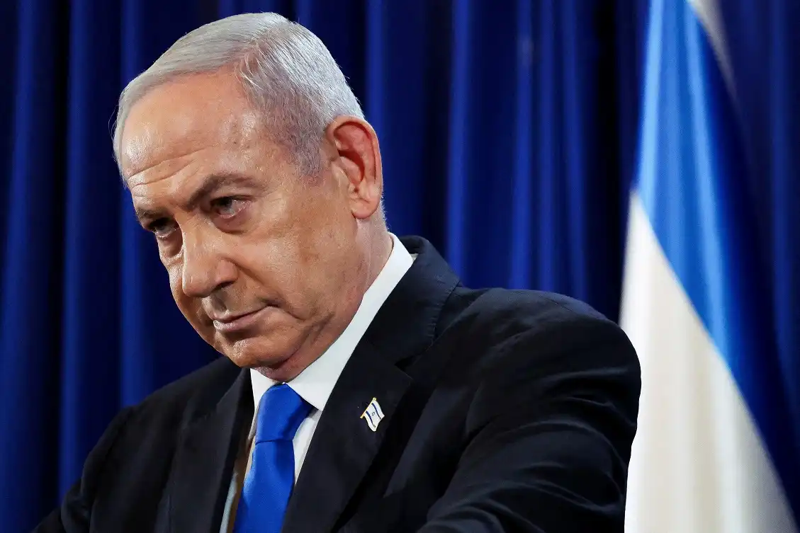 Benjamin Netanyahu em uma sala, olhando de baixo para cima, com cortinas azuis e a bandeira de Israel ao fundo, refletindo sobre os próximos passos políticos