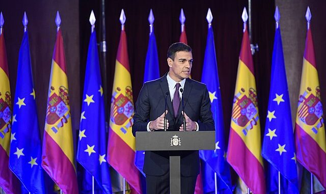 Pedro Sánchez discursando em conferência com bandeiras da Espanha e da União Europeia ao fundo.