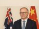 Primeiro-Ministro australiano Anthony Albanese em pé, vestindo terno escuro, gravata azul e óculos, com as bandeiras da Austrália e da China ao fundo.