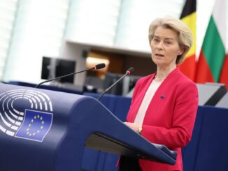 Presidente da Comissão Europeia falando no plenário do Parlamento Europeu, com bandeiras nacionais ao fundo e o símbolo da União Europeia no púlpito.