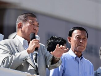 Dois homens japoneses, um deles falando ao microfone durante um pronunciamento público.