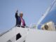 Donald Trump acenando com o polegar para cima ao embarcar em avião presidencial sob céu azul claro.