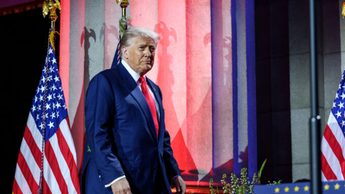 Donald Trump diante de colunas iluminadas em vermelho e azul, com bandeiras dos Estados Unidos ao fundo