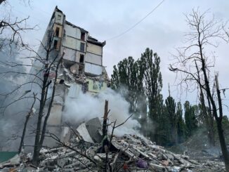 Prédio parcialmente destruído em Kiev após ataque aéreo russo, com fumaça subindo dos escombros e árvores danificadas ao redor.