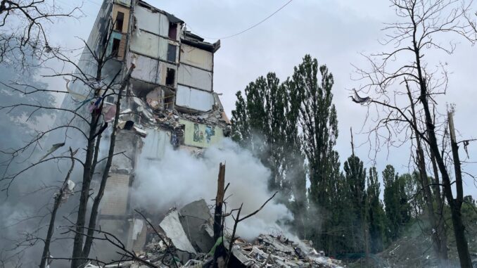 Prédio parcialmente destruído em Kiev após ataque aéreo russo, com fumaça subindo dos escombros e árvores danificadas ao redor.