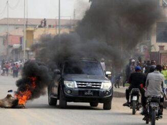 Carro preto atravessando rua com pneus em chamas e fumaça preta densa durante protestos em Luanda, com pessoas observando e motociclistas passando ao lado.