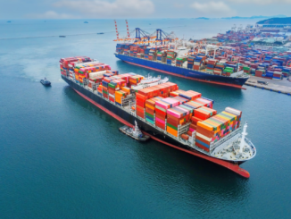 Navio porta-contêineres carregado com contêineres coloridos navegando em um porto movimentado com outros navios e guindastes ao fundo.