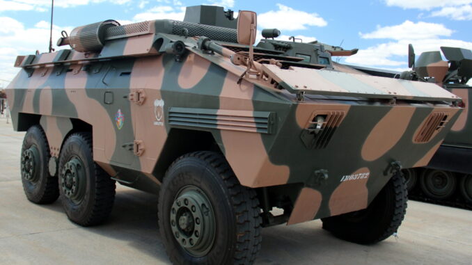 Veículo blindado EE-11 Urutu com camuflagem verde e marrom estacionado em área externa, sob céu azul com nuvens.
