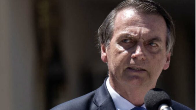 Jair Bolsonaro discursando em microfone, terno azul, expressão séria, em evento ao ar livre.