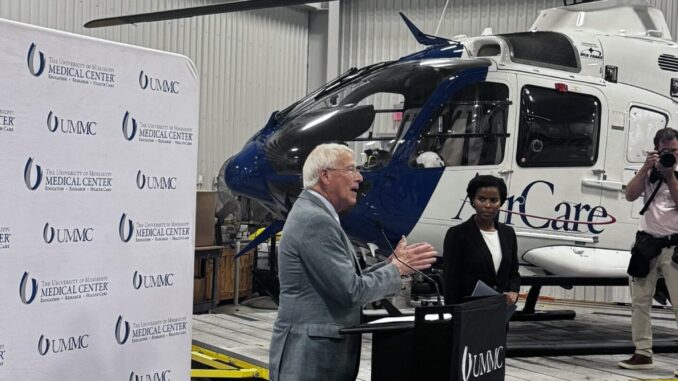 Senador Roger Wicker falando em um púlpito com logotipo da UMMC, com um helicóptero AirCare ao fundo e fotógrafos registrando o evento.