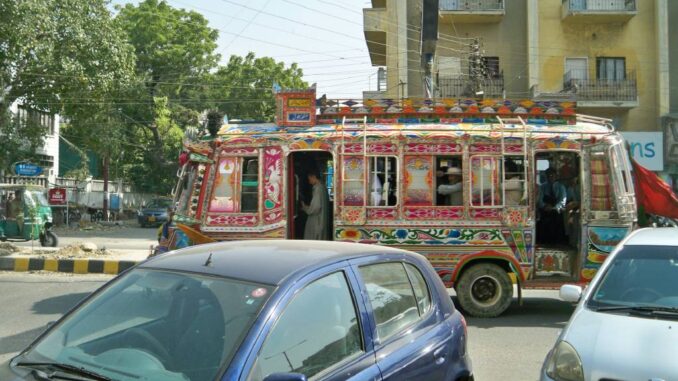 Ônibus decorado colorido circulando em rua urbana de Karachi, Paquistão, com carros e edifícios ao fundo.