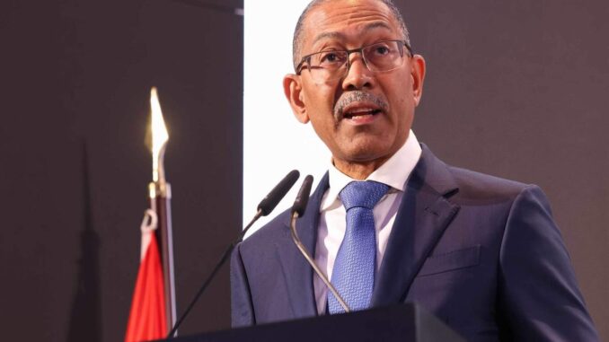 Ministro Diamantino Pedro Azevedo de Angola falando em um púlpito durante evento oficial, usando terno azul, gravata azul clara e óculos, com microfones à frente e bandeira de Angola ao fundo.