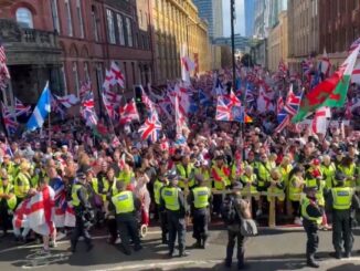 Multidão de manifestantes em Londres segurando bandeiras britânicas, cruzes de São Jorge e bandeiras de países estrangeiros, com policiais em coletes amarelos formando barreiras à frente.