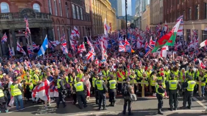 Multidão de manifestantes em Londres segurando bandeiras britânicas, cruzes de São Jorge e bandeiras de países estrangeiros, com policiais em coletes amarelos formando barreiras à frente.