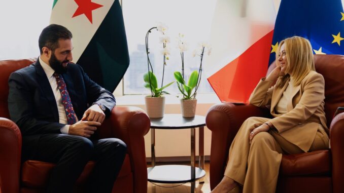 Encontro entre Ahmed al-Sharaa, presidente da Síria, e representante italiana durante a Semana de Alto Nível da Assembleia Geral da ONU, com bandeiras da Síria, Itália e União Europeia ao fundo.