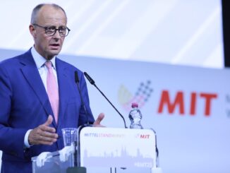 Friedrich Merz, chanceler alemão, discursando em um púlpito durante o evento MIT, usando terno azul e gravata rosa, com microfones à frente e um fundo com o logotipo do MIT.