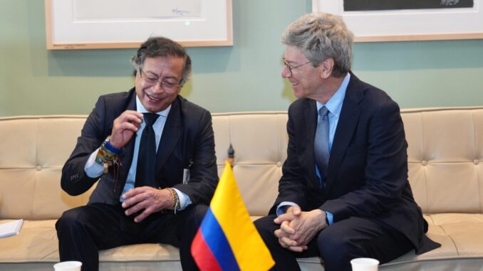 Gustavo Petro sentado à esquerda conversando com Jeffrey Sachs à direita, sobre integração econômica da América Latina, com bandeira da Colômbia à frente.