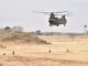 Helicóptero militar japonês transporta soldados por corda durante exercício no Acampamento Narashino, com tropas correndo no campo e outros helicópteros ao fundo.