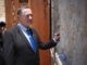 O ministro da Defesa de Israel, Israel Katz, vestido com terno preto e gravata azul, encosta a mão no Muro das Lamentações em Jerusalém, enquanto mensagens de papel estão inseridas entre as pedras.