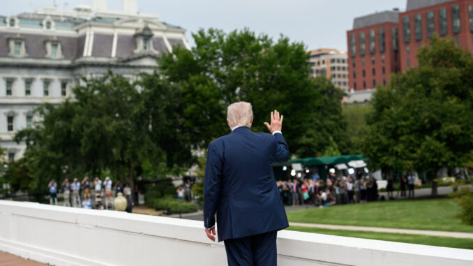 Presidente Donald Trump de costas, acenando para um grupo de jornalistas em frente a prédios governamentais em Washington, EUA.