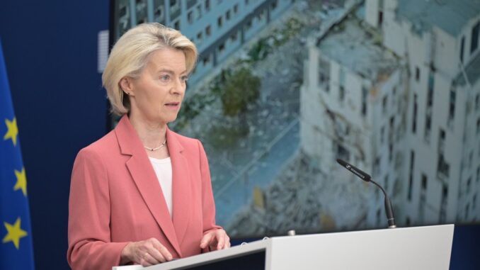 Ursula von der Leyen, presidente da Comissão Europeia, falando em frente a um microfone, com a bandeira da União Europeia ao fundo e imagens de destruição de prédios em tela atrás dela.