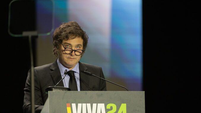 Javier Milei discursando em púlpito com microfone, usando óculos e terno, durante evento VIVA24.