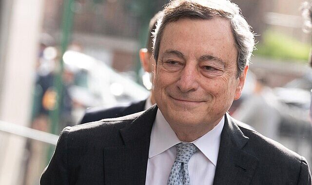 Mario Draghi sorrindo, vestindo terno escuro e gravata, em evento público ao ar livre.