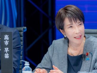 Sanae Takaichi, candidata à liderança do Partido Liberal Democrata, sentada à mesa de debate, gesticulando enquanto fala, vestindo blazer cinza e microfone preso à roupa, durante evento político em 28 de setembro de 2025.