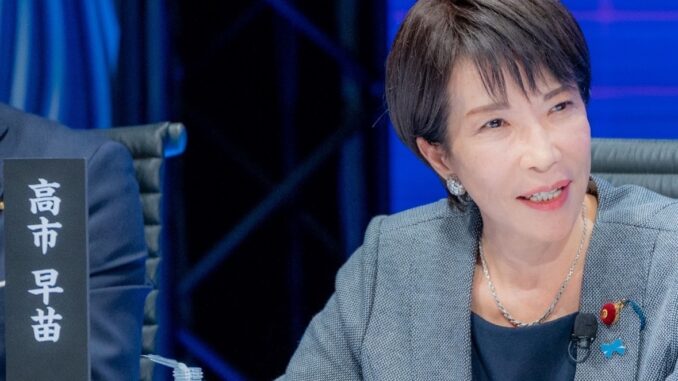 Sanae Takaichi, candidata à liderança do Partido Liberal Democrata, sentada à mesa de debate, gesticulando enquanto fala, vestindo blazer cinza e microfone preso à roupa, durante evento político em 28 de setembro de 2025.