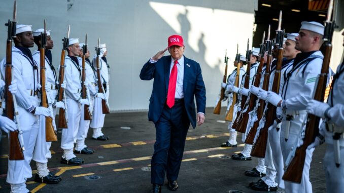 Donald Trump, usando boné vermelho com "USA", saudando tropas dos EUA em formação militar, com fuzis e uniformes brancos.