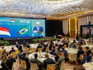 Momento do Fórum Empresarial Indonésia-Brasil com participantes sentados à mesa e um orador no palco, com as bandeiras do Brasil e da Indonésia ao fundo.