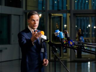 Rutte fala à imprensa durante coletiva, com múltiplos microfones de diferentes veículos de mídia, em ambiente moderno com portas de vidro ao fundo.