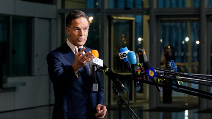 Rutte fala à imprensa durante coletiva, com múltiplos microfones de diferentes veículos de mídia, em ambiente moderno com portas de vidro ao fundo.