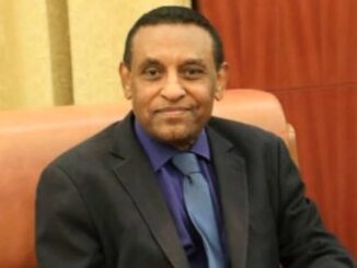 Fotografia de Kamil Idris, atual primeiro-ministro do Sudão, sentado em um sofá, vestindo terno escuro, camisa azul e gravata azul clara, olhando para a câmera com leve sorriso.