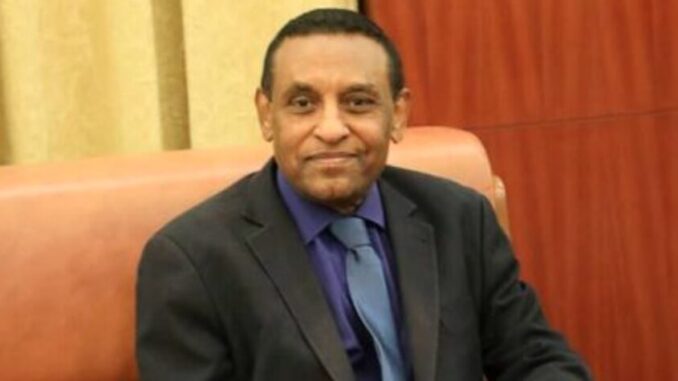 Fotografia de Kamil Idris, atual primeiro-ministro do Sudão, sentado em um sofá, vestindo terno escuro, camisa azul e gravata azul clara, olhando para a câmera com leve sorriso.