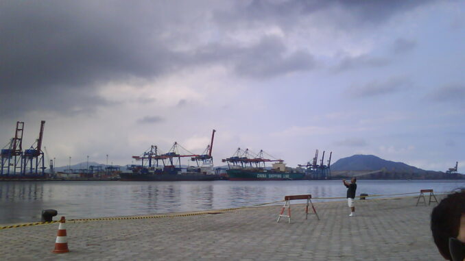 Vista panorâmica do Porto de Santos mostrando navios de carga e guindastes em operação, com céu nublado e infraestrutura portuária em destaque.