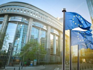 Bandeira da União Europeia em frente ao Parlamento Europeu em Bruxelas, com edifício moderno ao fundo.
