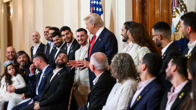 O presidente Donald Trump conversa sorrindo com ex-reféns israelenses libertados durante encontro oficial na Casa Branca, com bandeira dos Estados Unidos ao fundo.