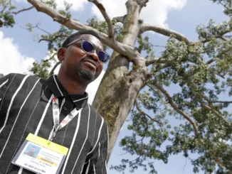 Olumide Idowu, ativista climático nigeriano, usa crachá de observador enquanto caminha ao ar livre próximo a uma grande árvore em Belém (PA).