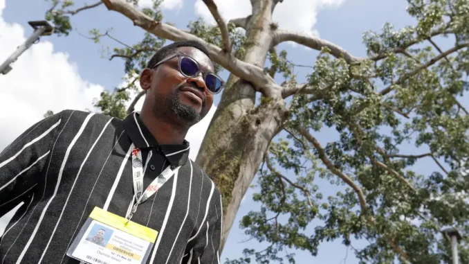 Olumide Idowu, ativista climático nigeriano, usa crachá de observador enquanto caminha ao ar livre próximo a uma grande árvore em Belém (PA).